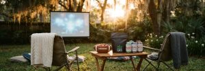 An editorial lifestyle photo of a Central Florida backyard game day setup during golden hour. In the background, a portable projector screen glows softly with an out-of-focus image. In the foreground, a low wooden table sits between two folding chairs draped with team-neutral blankets; the table holds a football-shaped bowl of snacks, a matte black Leafy8 pouch, and a cluster of Papa's Alternative Brew cans. The scene features warm autumn lighting, a shallow depth of field, and a candid, film-shot aesthetic.