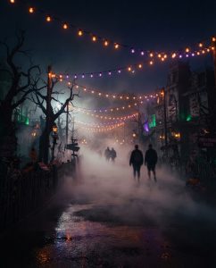 A moody nighttime path at a Halloween theme park event with rolling ground fog, overhead strings of orange and purple lights, and silhouettes of haunted house facades.