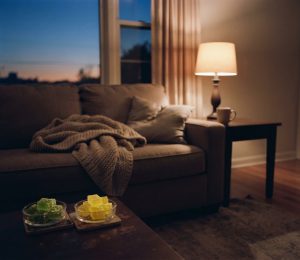 A candid, warm-toned evening photograph of a cozy living room scene. In the foreground, on a coffee table, rest two glass dishes: one containing vibrant Leafy8 relax gummies and the other containing Leafy8 sleep gummies. The midground features a comfortable sofa draped with a textured knit blanket and a plush pillow. To the right, a dark wood side table holds a glowing table lamp and a ceramic mug. In the background, a large window reveals a peaceful twilight gradient of deep blue and orange. No people are present. The overall mood is calm and restful.