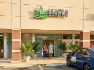 A two-person couple walking out of a Leafy8 retail hemp store in Central Florida during warm afternoon light, with green potted palms flanking the open glass doorway and a blue delivery sedan parked out front.