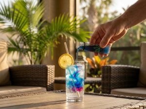 A lifestyle photo of a hand pouring blue syrup from a small Leafy8 bottle into a tall glass of sparkling water with ice and a lemon wheel on an outdoor patio. 